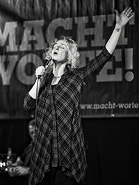 Bühnenfoto von Monika Mertens in schwarz-weiß bei einem Poetry Slam, performt leidenschaftlich mit starken Gesten auf der Bühne.